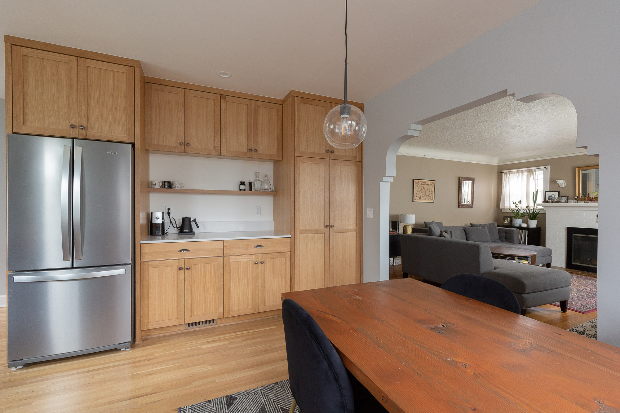 Pantry wall with refrigerator and coffee station, dining table in foreground, and living room visible through the reconstructed decorative archway beyond, Arbor Lodge Kitchen by Tweto Architecture
