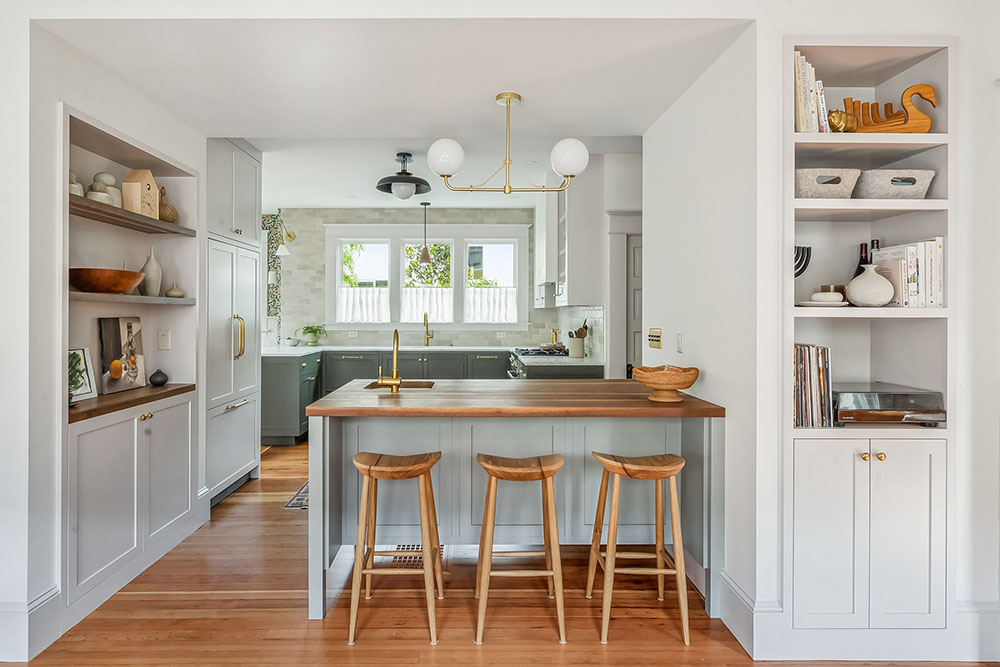 Remodeled farmhouse kitchen with sage cabinets, a walnut-topped island with stools, and flanking built-in shelves, Baldwin Street Remodel by Tweto Architecture, North Portland