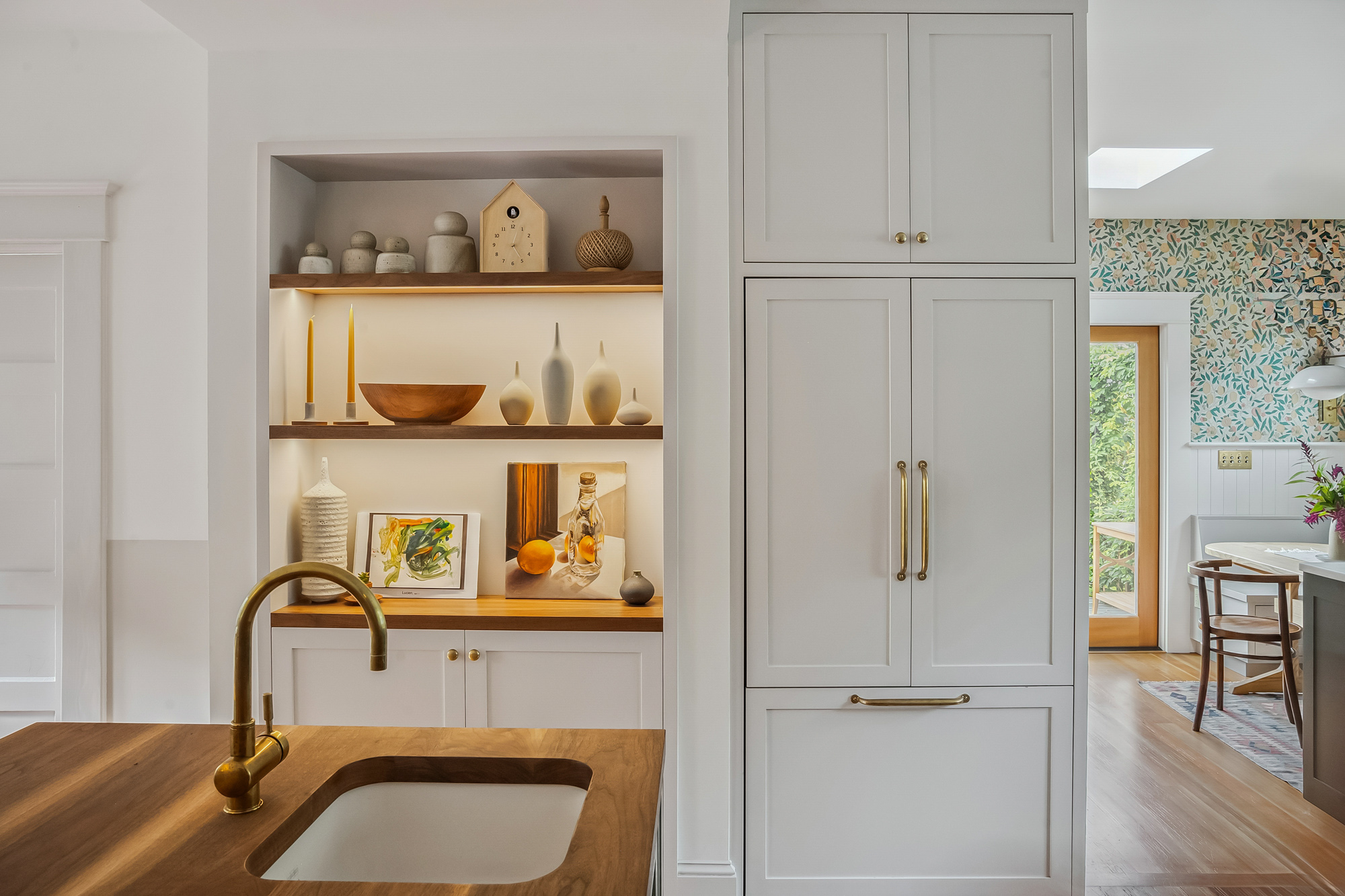 Lit built-in display niche with walnut shelves beside the tall painted pantry, island with walnut countertop and brass faucet in the foreground, Baldwin Street remodel by Tweto Architecture