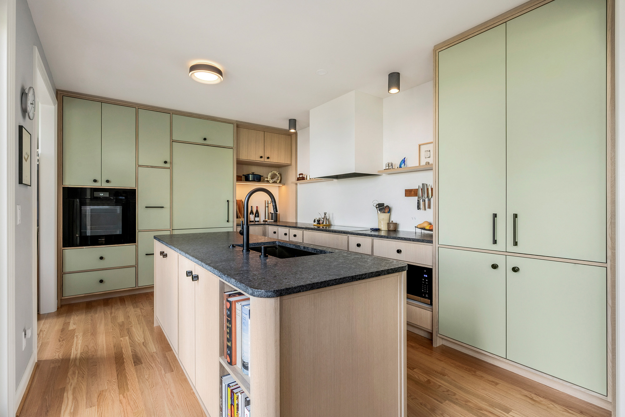 Kitchen with sage green linoleum cabinet fronts and natural oak carcasses, oak island with leathered granite and open cookbook shelving at end, by Tweto Architecture