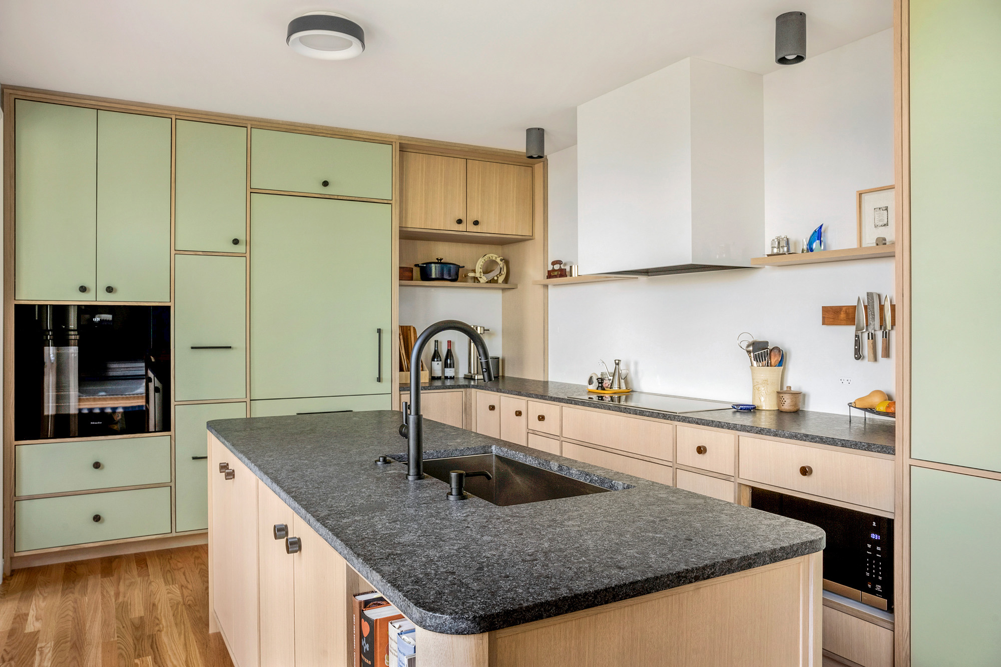 Island from kitchen side showing leathered granite counter with integrated black sink, sage tall cabinet wall and open shelves behind, by Tweto Architecture