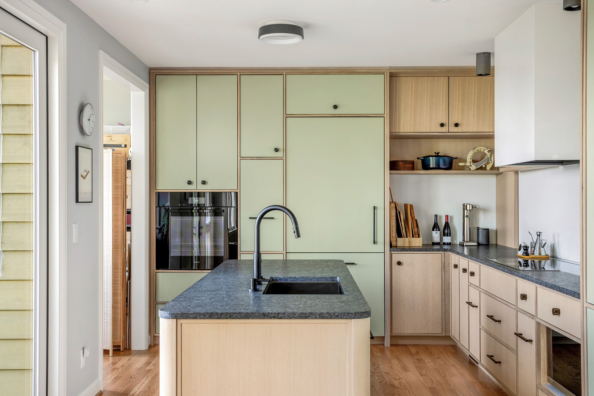 Kitchen from entry side showing full tall cabinet wall with integrated appliances, island with sink in foreground, terrace door visible at rear, by Tweto Architecture