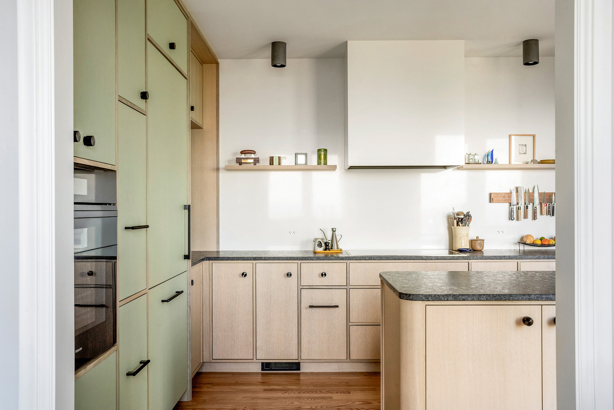 Cooking side of kitchen with induction cooktop, open floating shelves, and sage tall cabinet wall with appliances at left, by Tweto Architecture