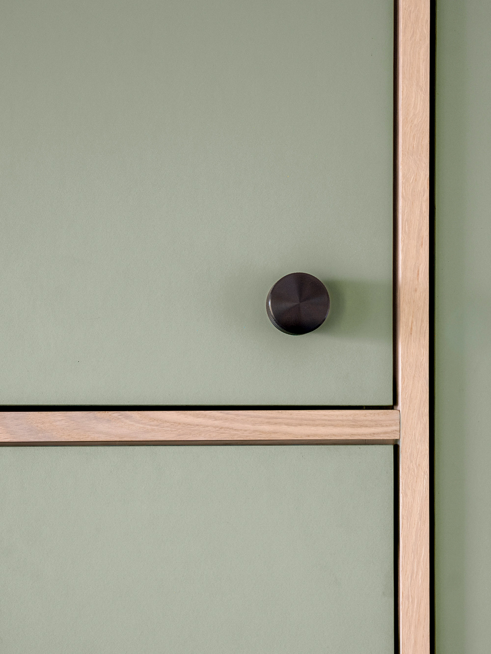 Close-up of sage linoleum cabinet door face with round black hardware knob and oak frame, by Tweto Architecture