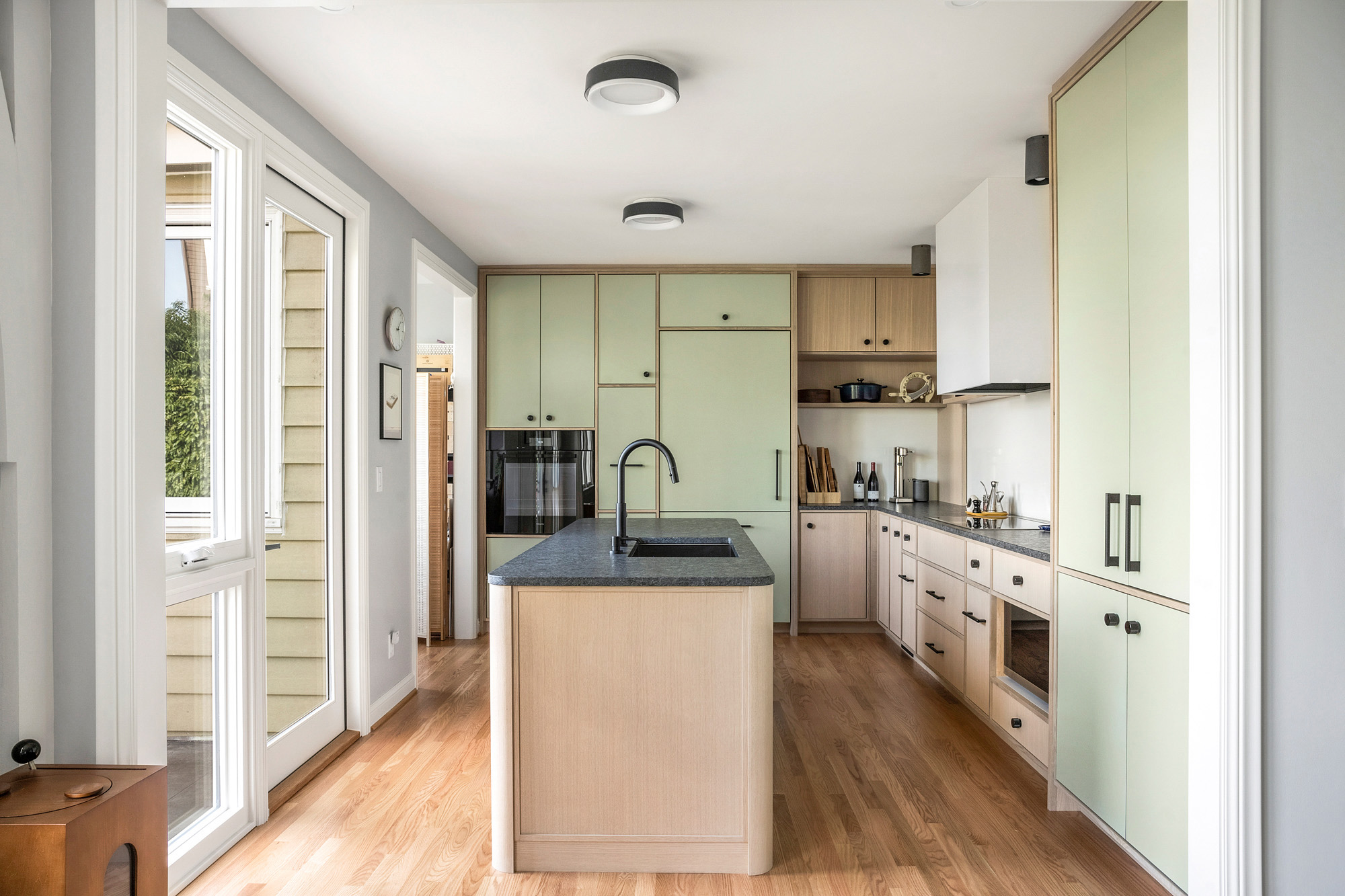 Remodeled kitchen with sage cabinetry, island with sink, and full-height terrace doors opening to deck and west hills views, by Tweto Architecture
