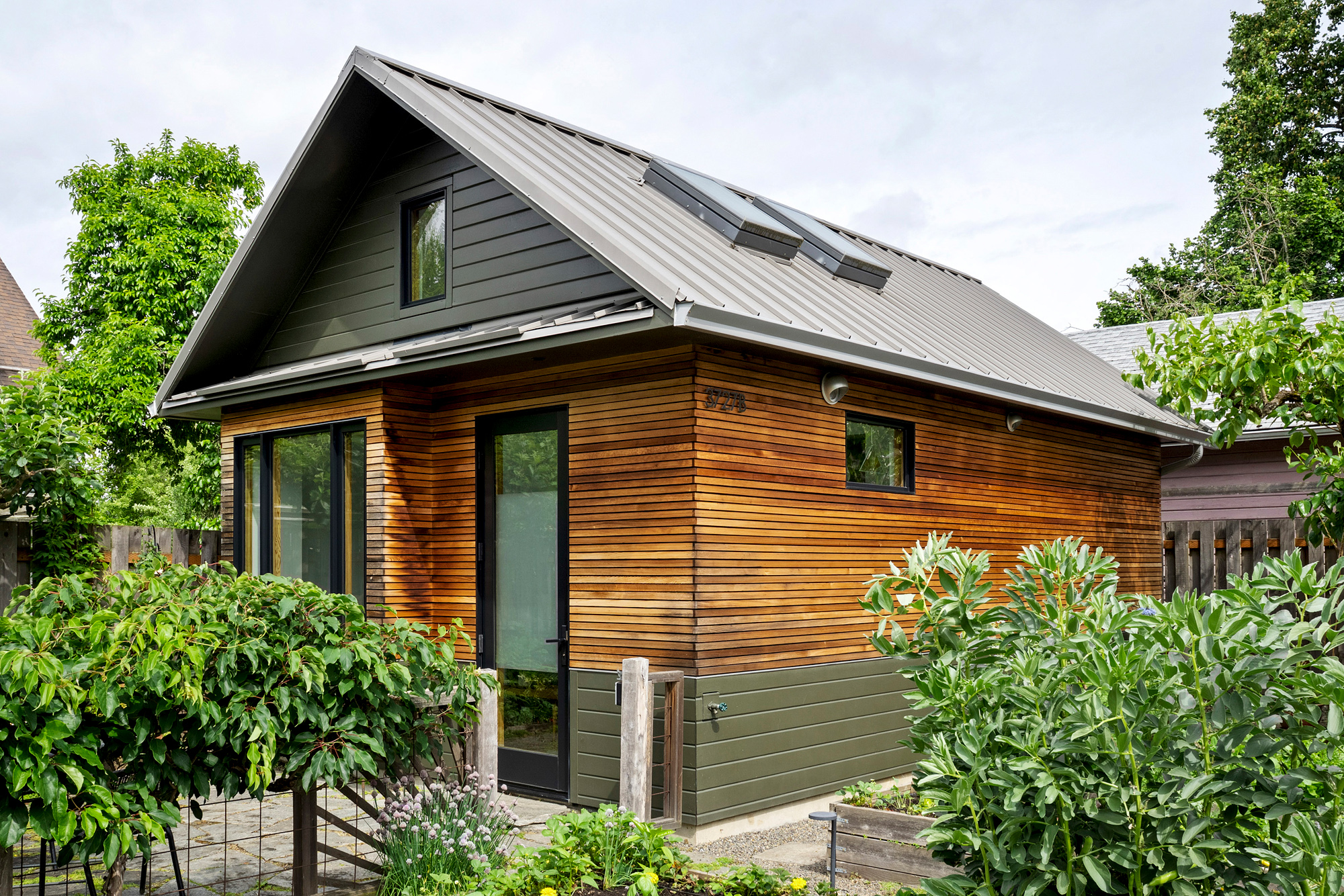 Front corner of the Irving Park ADU showing cedar lap siding, full-height glazed doors and windows, and a standing-seam metal roof with skylights, surrounded by garden plantings, by Tweto Architecture