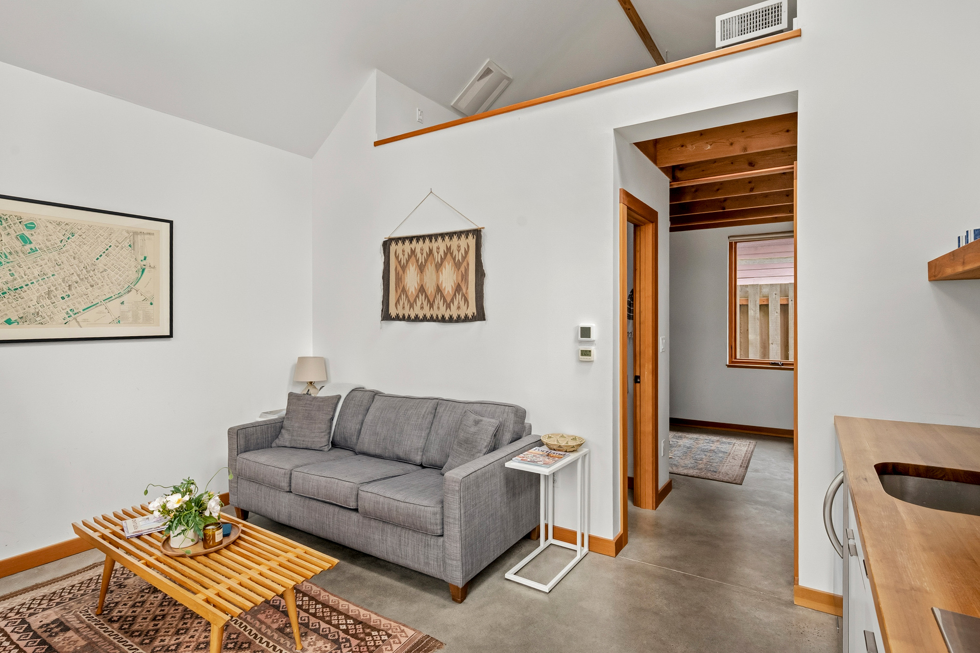 Living area with grey sofa, woven wall hanging, and polished concrete floor, with an open loft above and the bedroom visible through the doorway, Irving Park ADU by Tweto Architecture