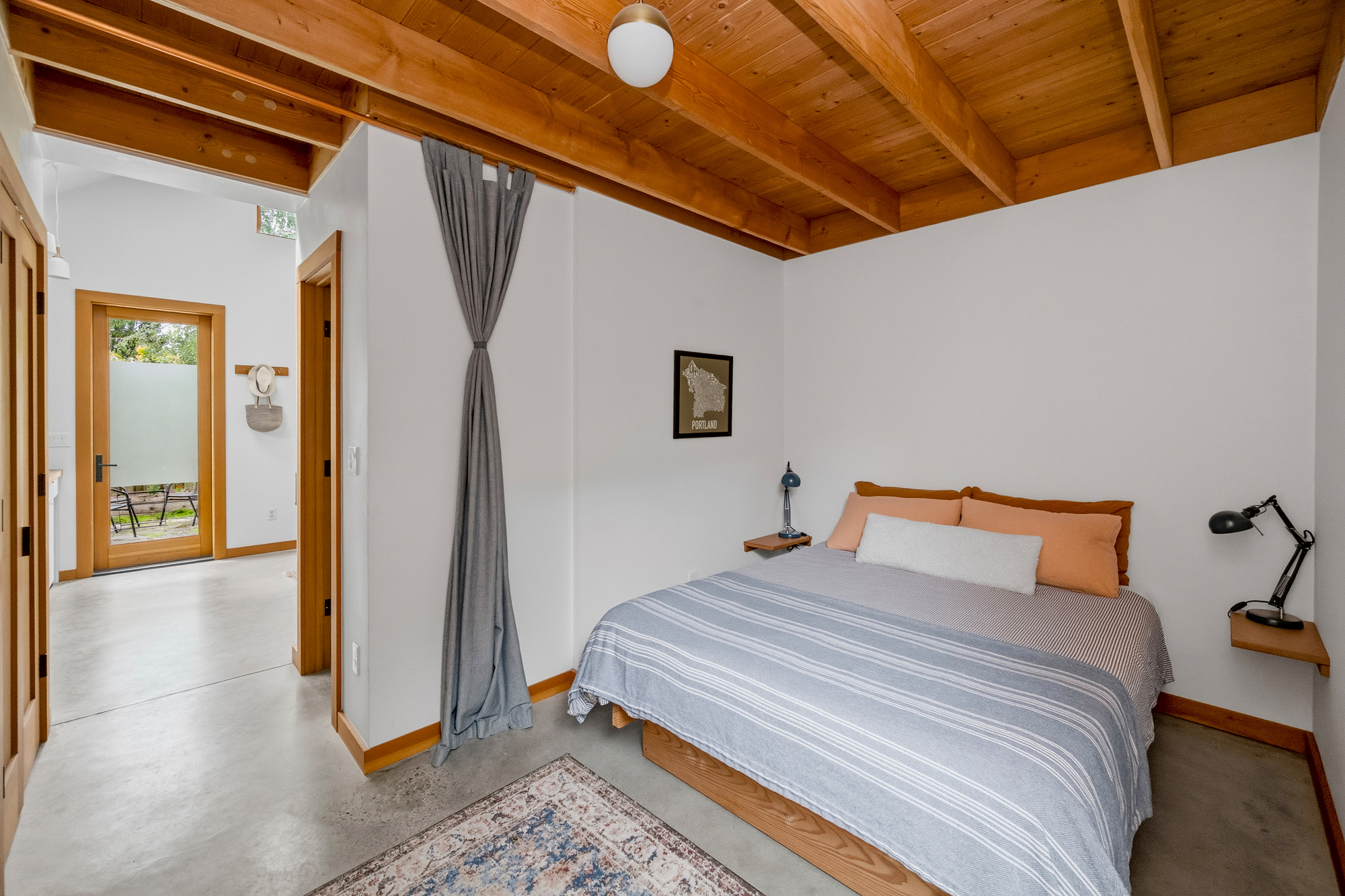 Bedroom with exposed wood ceiling and beams, looking out through the doorway toward the living area and glazed exterior door, Irving Park ADU by Tweto Architecture