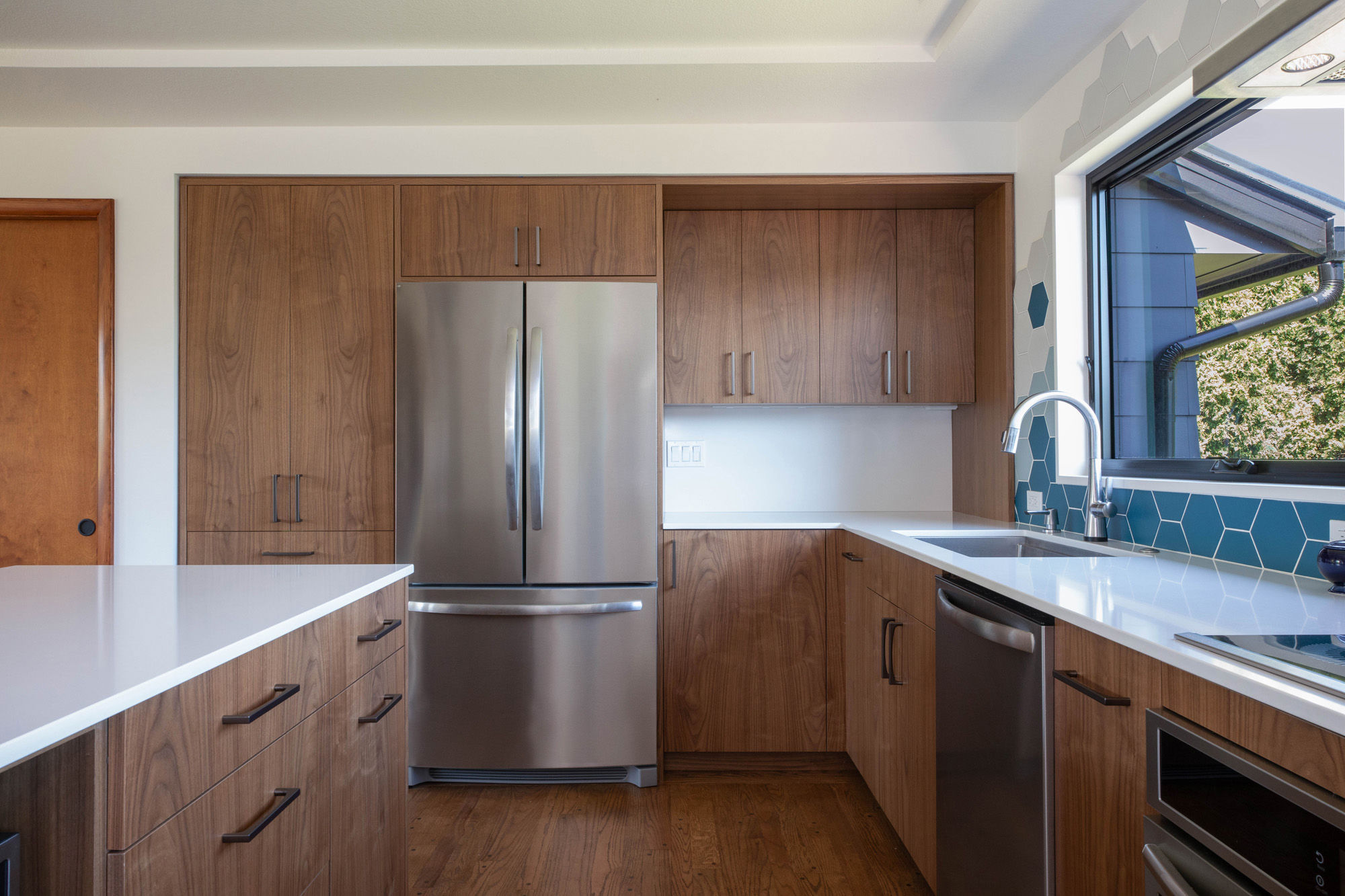 Kitchen showing walnut cabinetry, white quartz countertops, stainless refrigerator, and blue hexagonal tile backsplash with window over the sink, Kelly Street remodel by Tweto Architecture