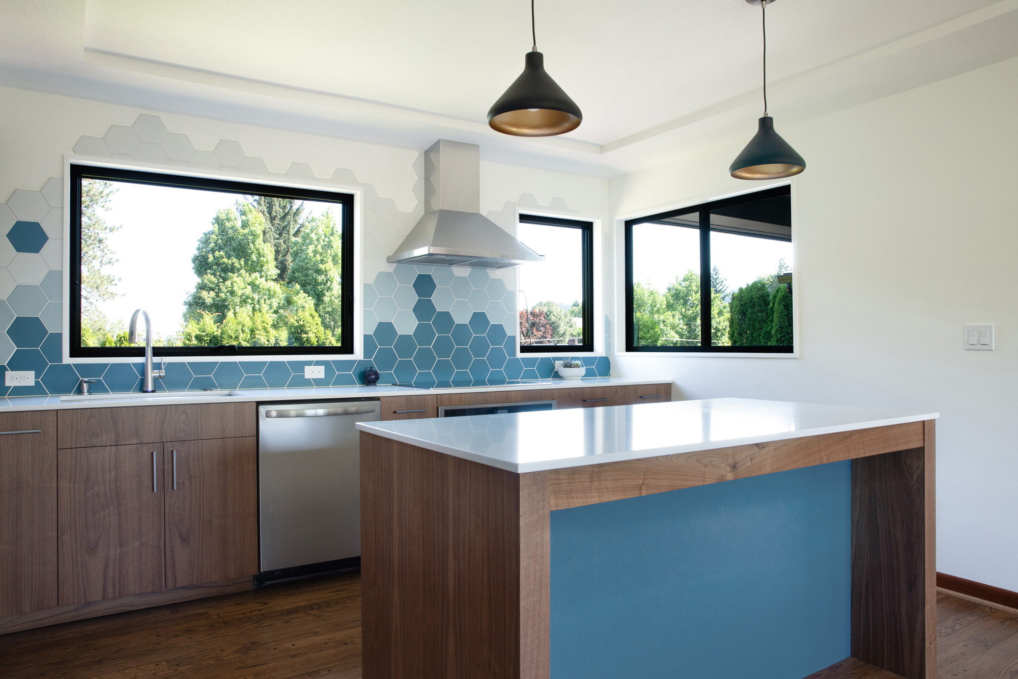 Kitchen island with blue end panel and walnut base, blue hexagonal tile backsplash wrapping two large backyard windows, black pendant lights above, Kelly Street remodel by Tweto Architecture