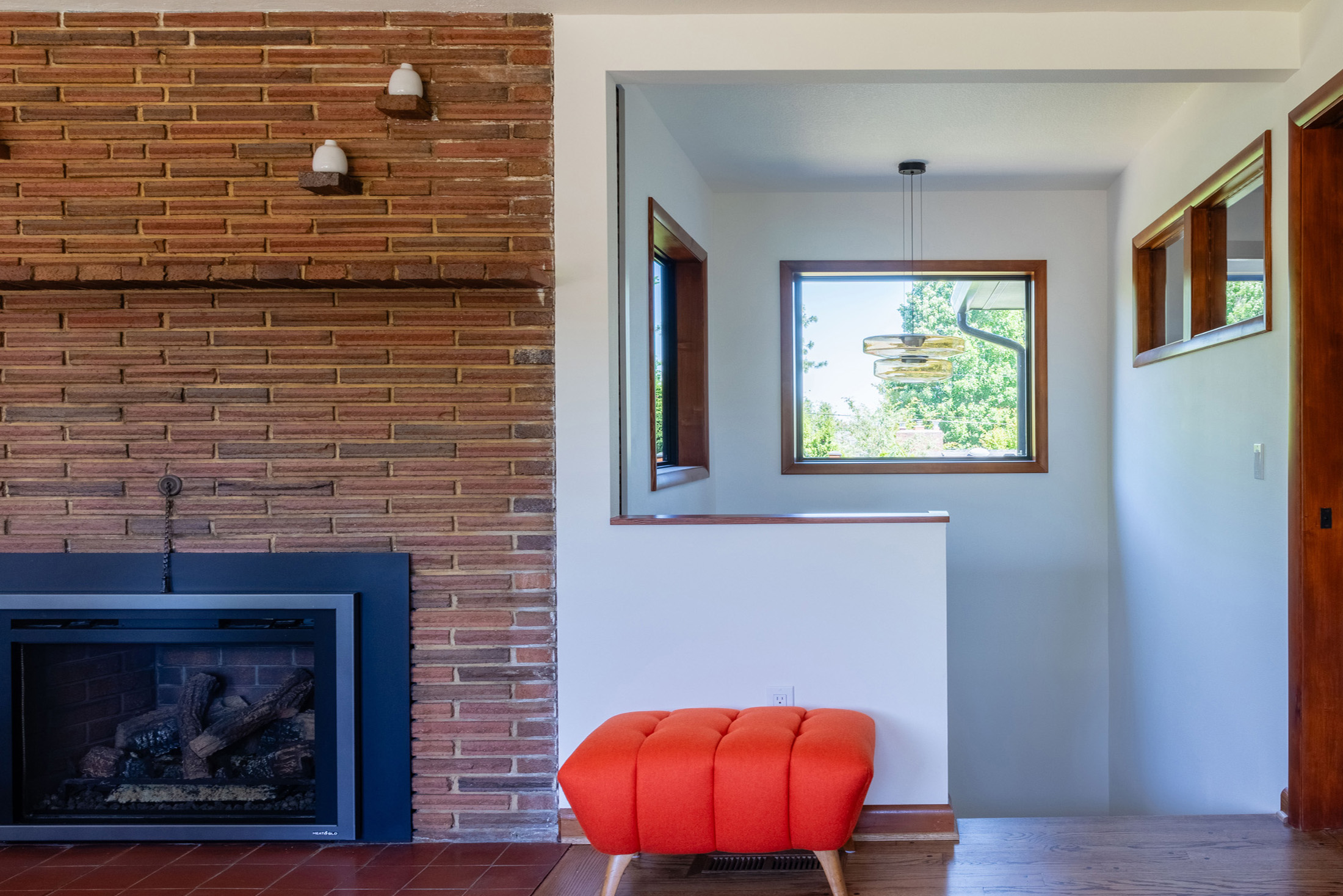 Living room with original brick fireplace, new stair opening cut through into the addition visible in the background, red ottoman in the foreground, Kelly Street remodel by Tweto Architecture