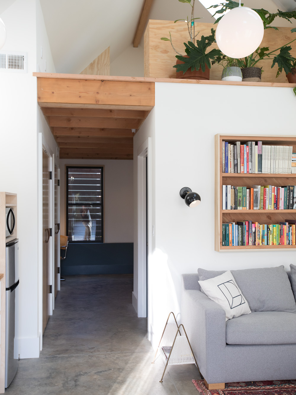 View from living room into the bedroom hallway with exposed fir ceiling, loft above with plants visible, built-in bookcase on right, Mason Street ADU by Tweto Architecture