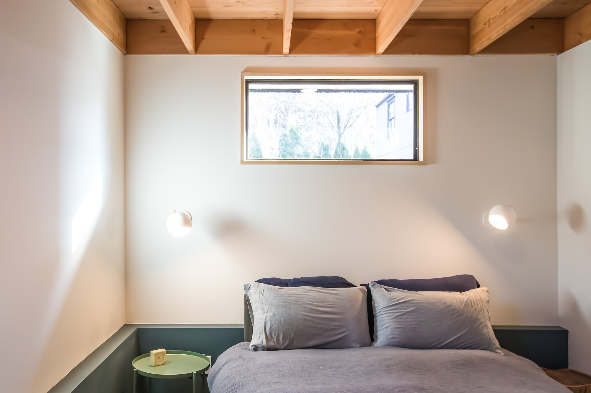 Bedroom with exposed Douglas Fir beam-and-deck ceiling, horizontal band window above the bed, and wall sconces on either side, Mason Street ADU by Tweto Architecture
