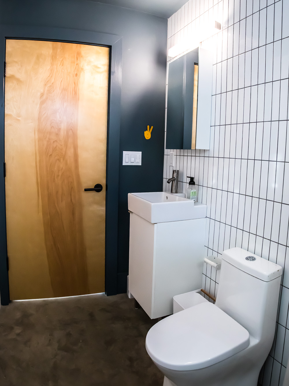 Bathroom with white square tile, pedestal vanity, and plywood door on dark teal walls, Mason Street ADU by Tweto Architecture