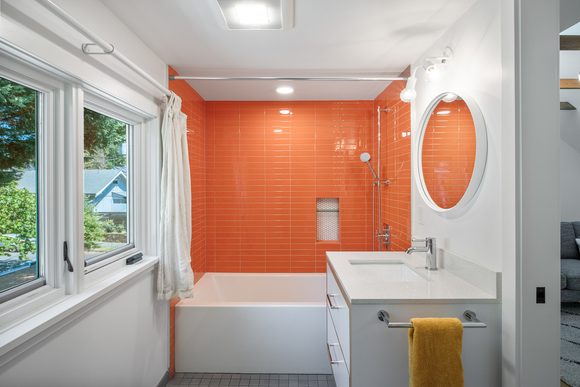 Attic bathroom with floor-to-ceiling orange tile on the tub surround, white vanity, and round mirror, NE 9th Remodel by Tweto Architecture