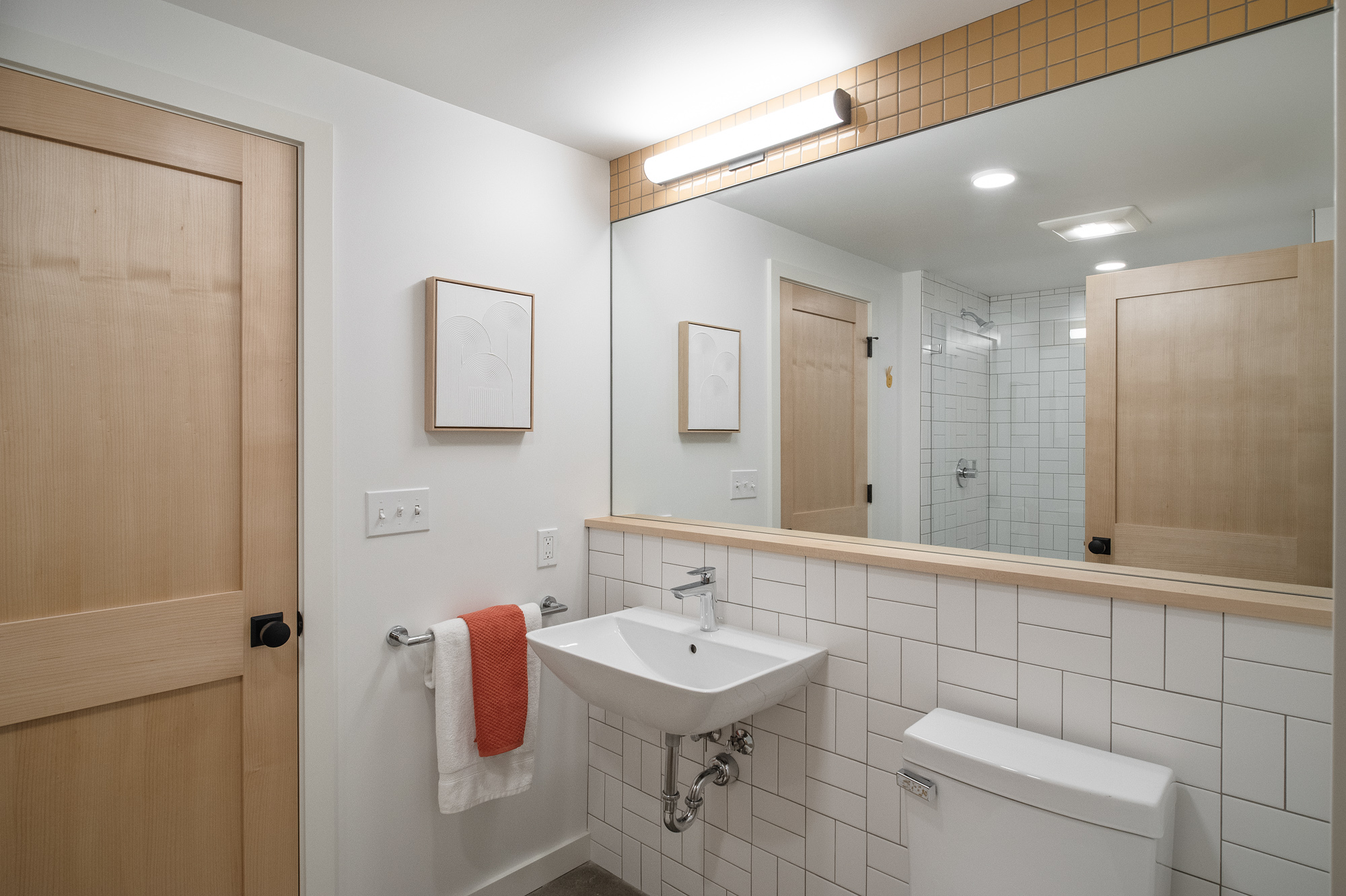 Basement bathroom with wall-mount sink, white subway tile, amber tile accent strip above the mirror, and glass shower visible in reflection, NE 9th Remodel by Tweto Architecture