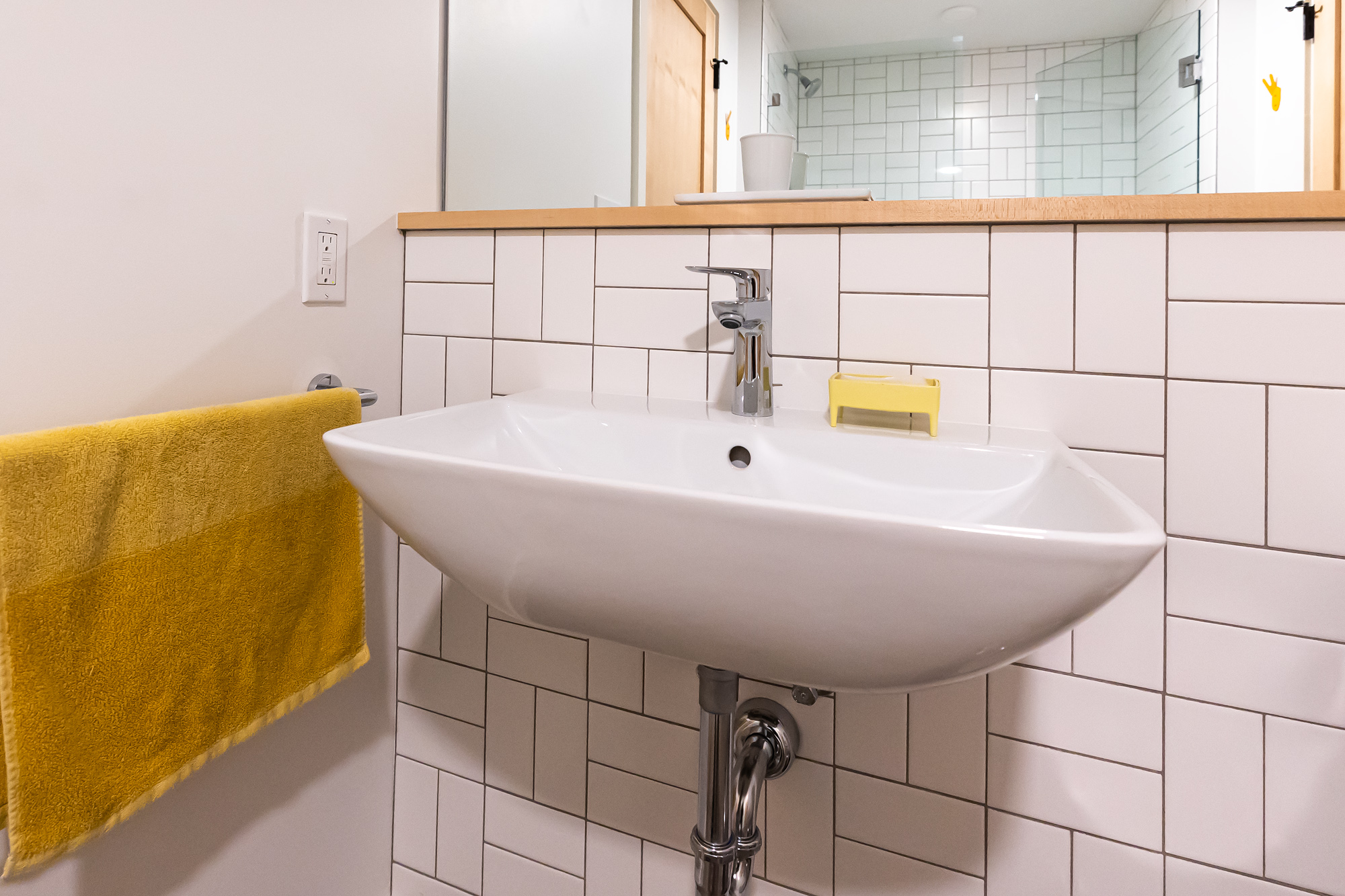 Close-up of wall-mount sink against white subway tile with maple counter ledge, glass shower visible in mirror reflection above, NE 9th Remodel by Tweto Architecture