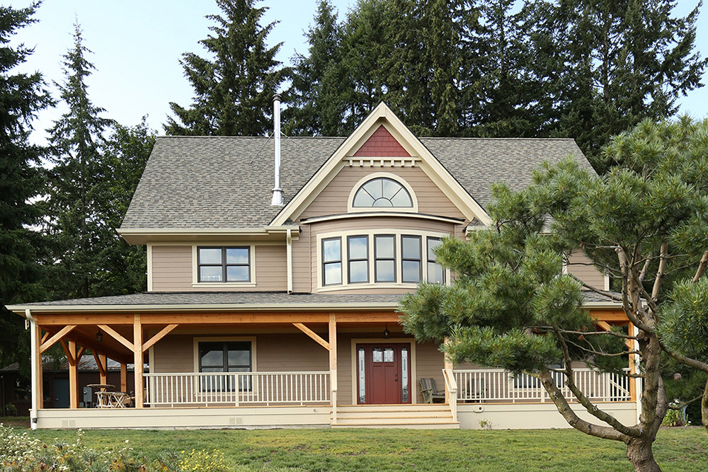 South-facing farmhouse exterior with wraparound timber porch, farmhouse gable with half-round window, and mature pine trees, North Plains Timber Frame by Tweto Architecture