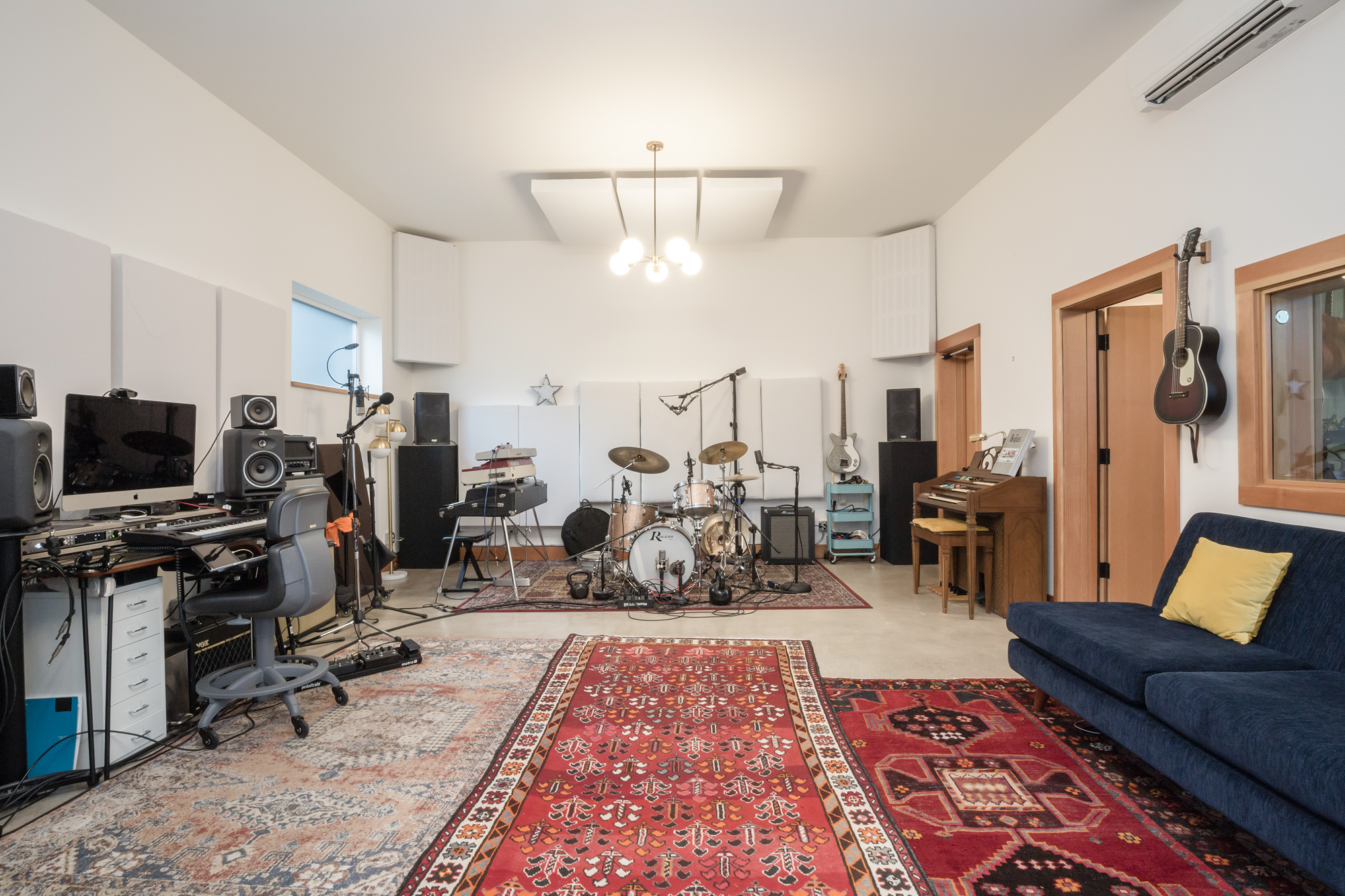 Live room with drum kit, mixing station, guitars on the wall, blue sofa, and tall acoustic panels below 11-foot ceilings, Siskiyou Street Recording Studio by Tweto Architecture