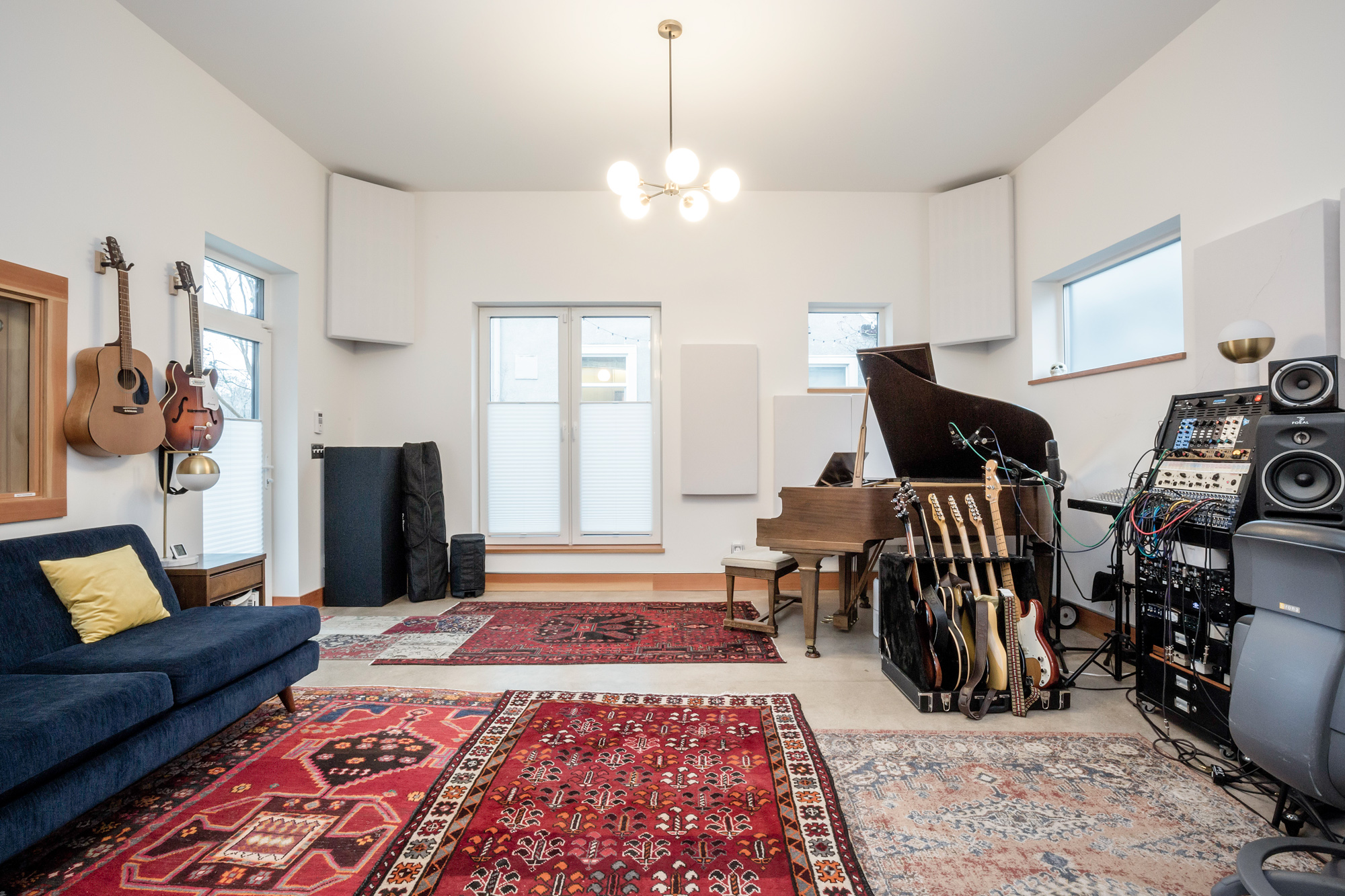 Live room from the opposite end showing grand piano, guitars on wall, blue sofa with yellow pillow, acoustic panels, and clerestory windows, Siskiyou Street Recording Studio by Tweto Architecture