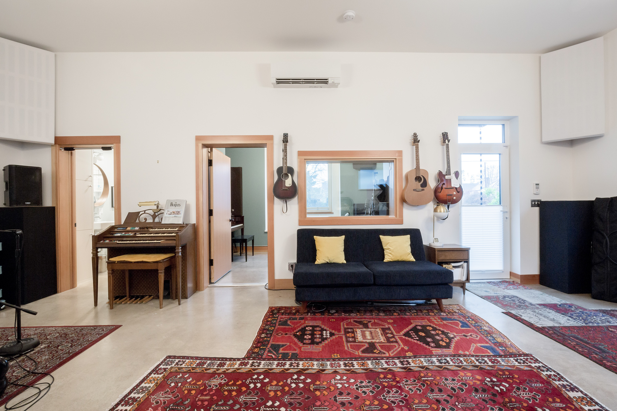 Live room looking toward the control room window with navy sofa and yellow pillows, guitars on the wall, and organ in the corner, Siskiyou Street Recording Studio by Tweto Architecture