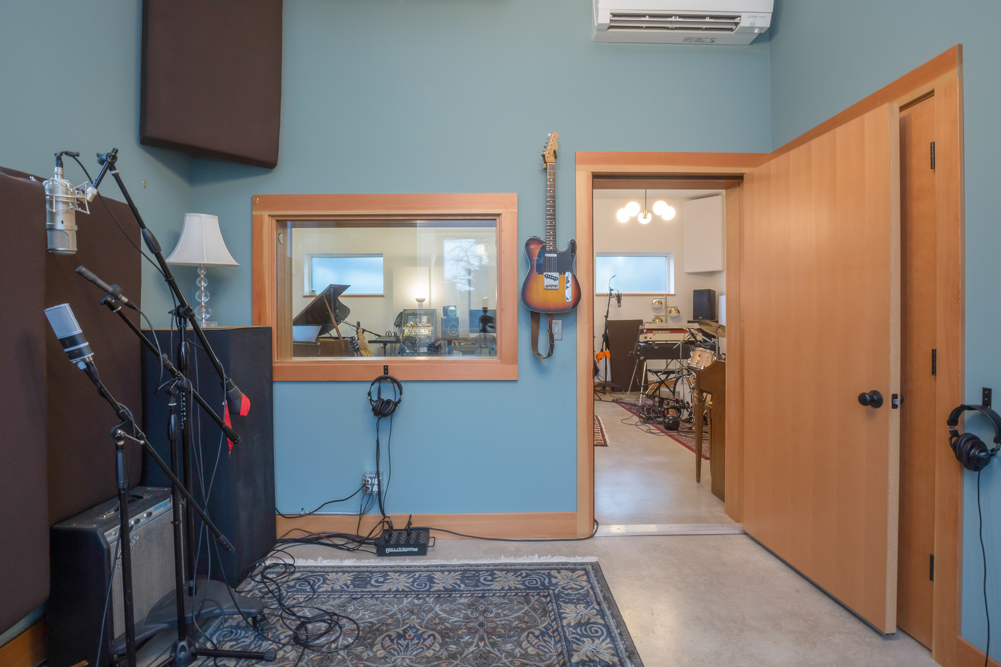 Control room with blue walls and interior glass window looking through to the live room, microphone stands and guitar in foreground, Siskiyou Street Recording Studio by Tweto Architecture