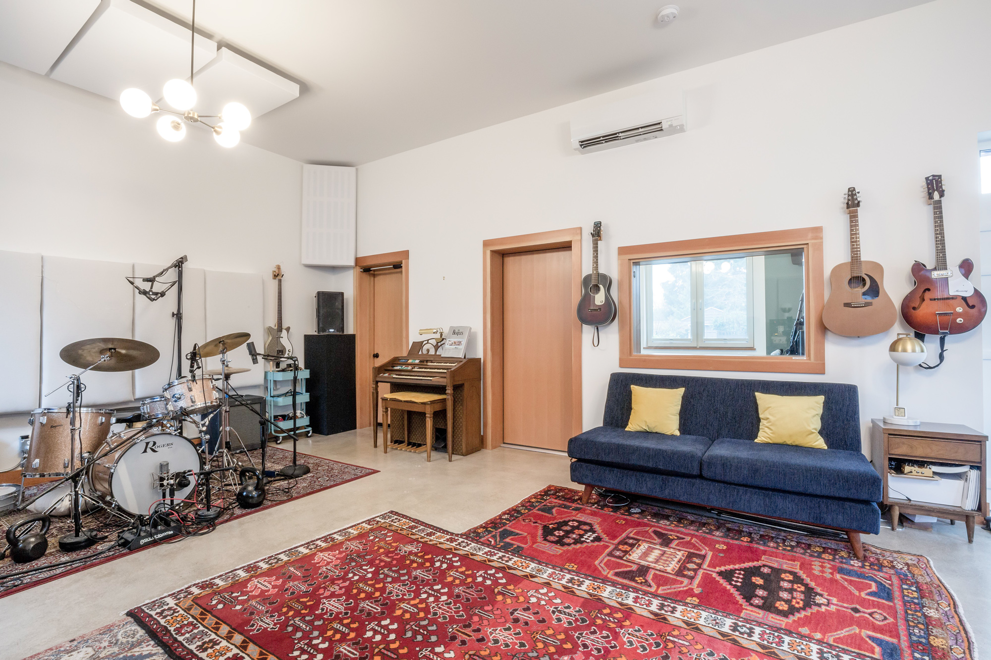 Live room with drum kit at left, navy sofa in the center, guitars and control room window at right, and chandelier above, Siskiyou Street Recording Studio by Tweto Architecture