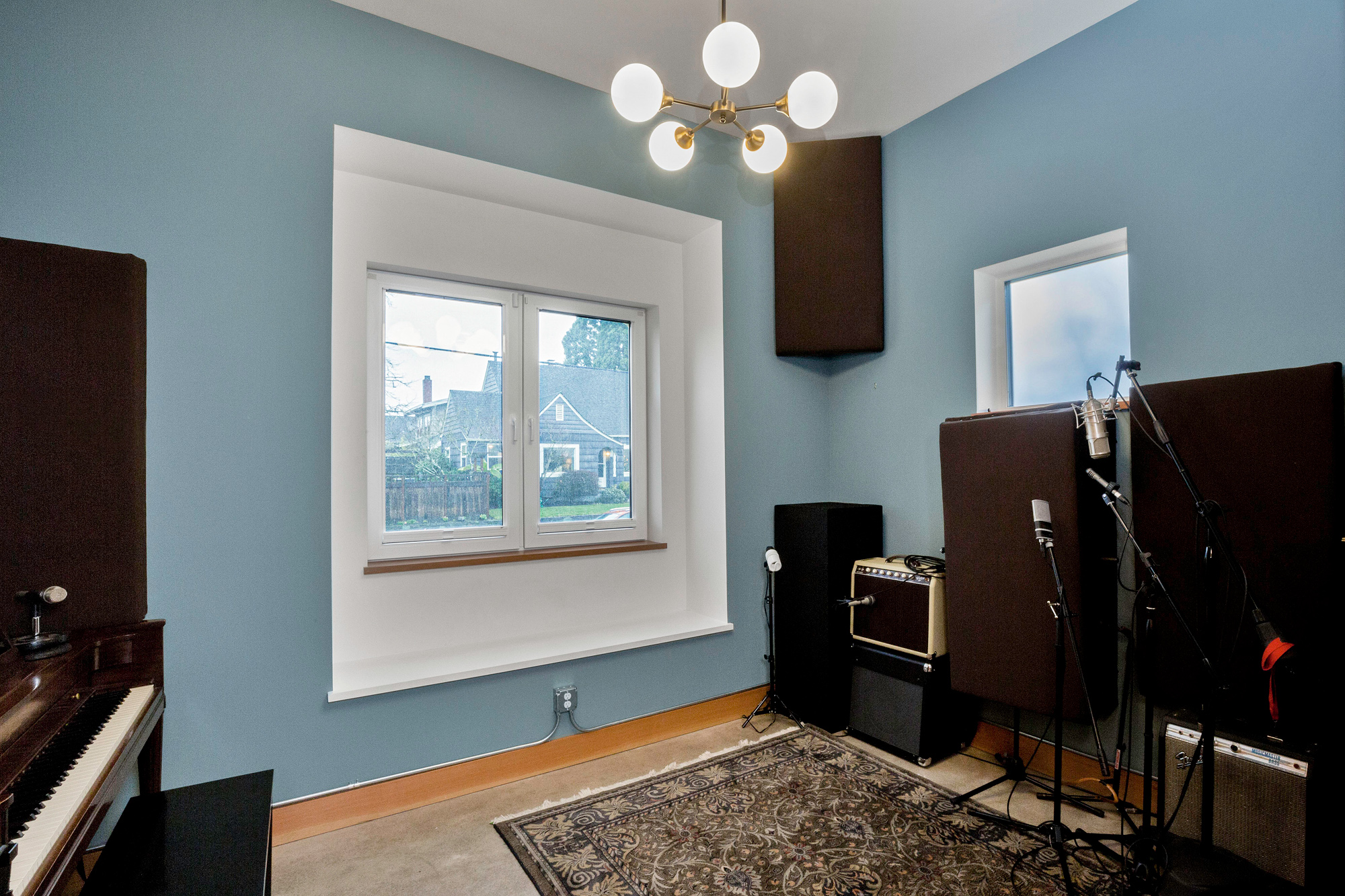 Control room with blue-gray walls and floating bay window to the street, microphone stands, guitar amp, and upright piano at left, Siskiyou Street Recording Studio by Tweto Architecture