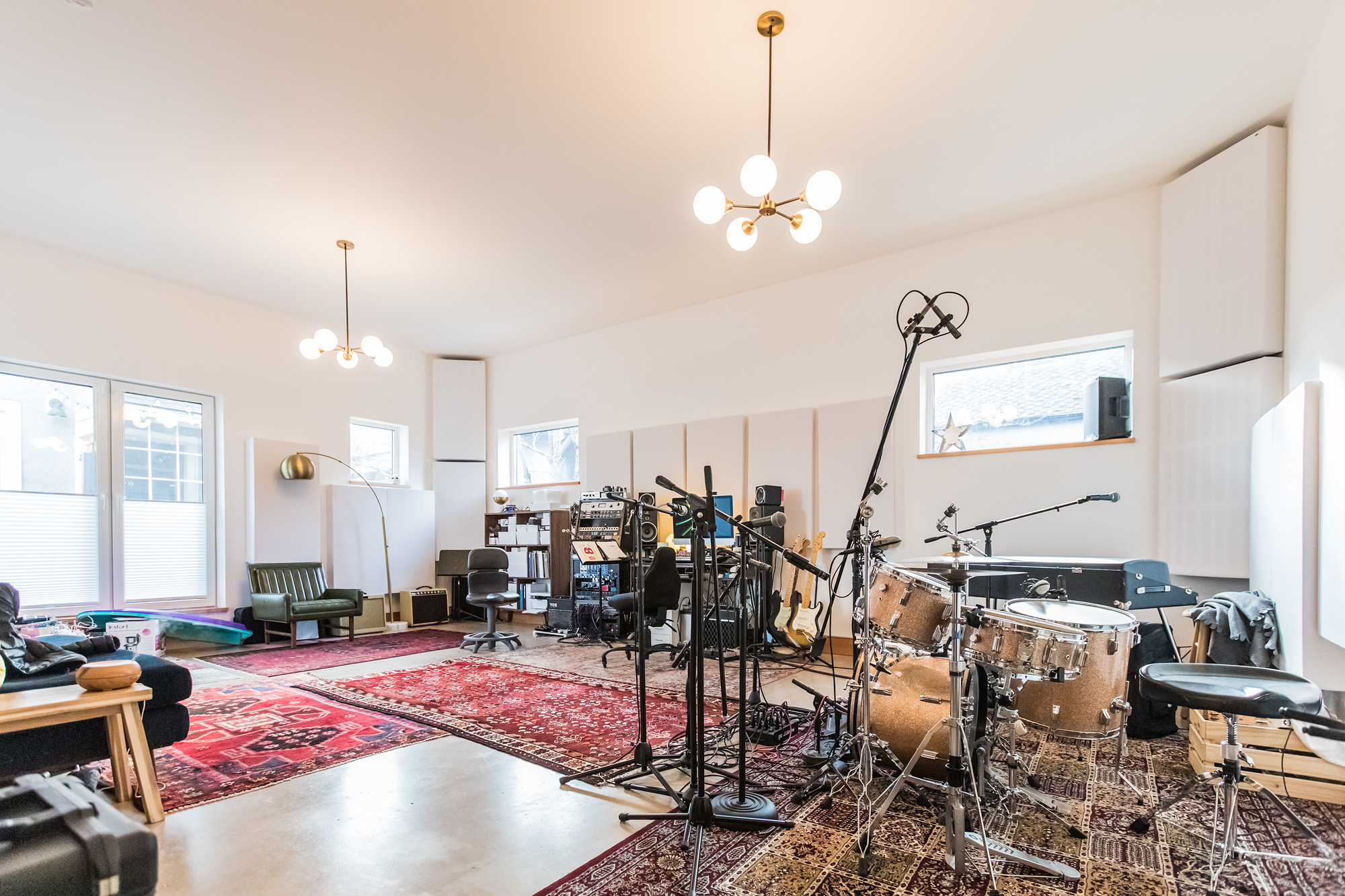 Full-length view of the live room with drum kit in foreground, two brass chandeliers, angled acoustic panels lining the tall walls, and control room visible at the far end, Siskiyou Street Recording Studio by Tweto Architecture