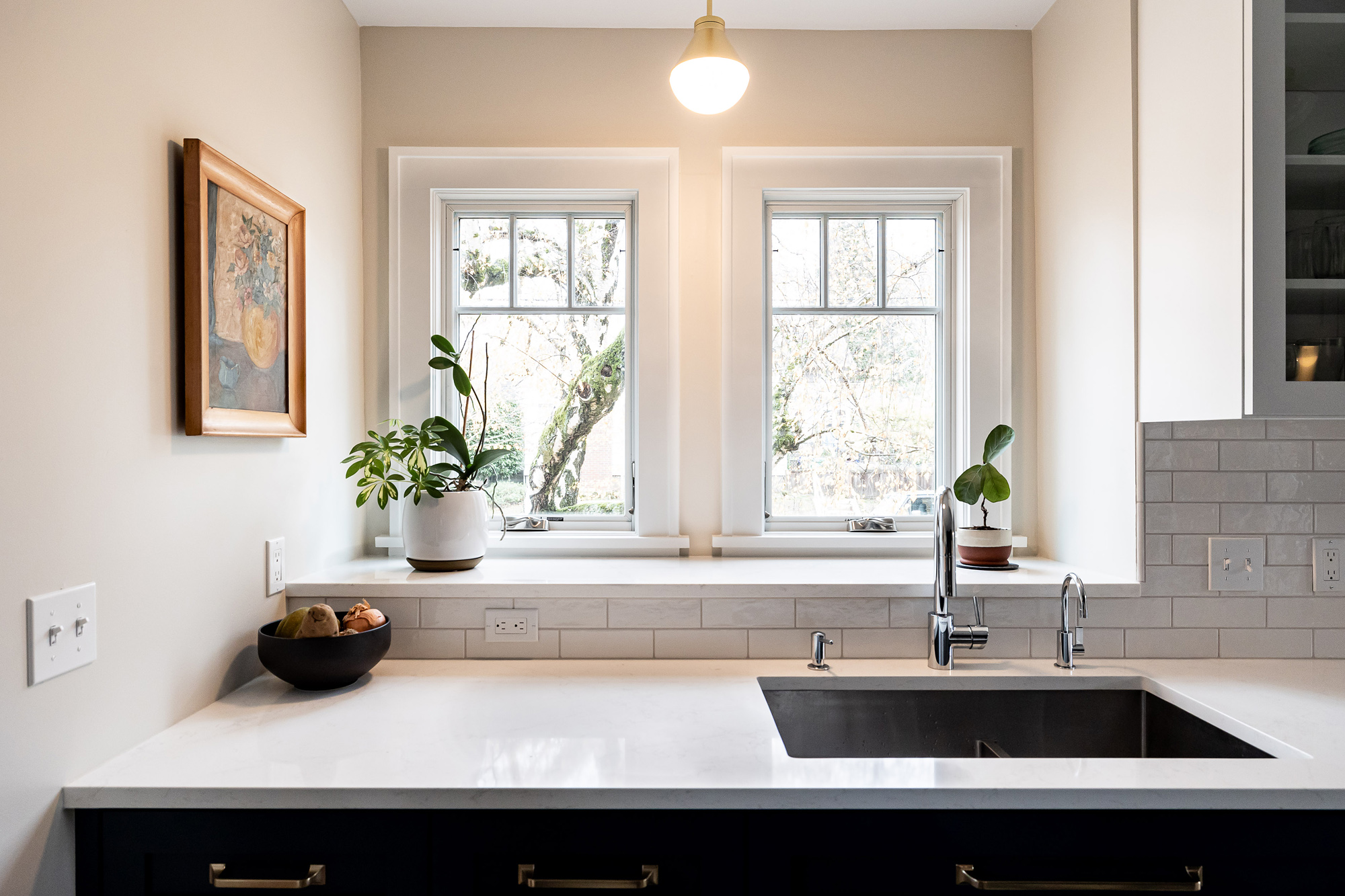 Kitchen sink with extra-deep window sill between two windows, quartz countertop, grey subway tile backsplash, and plants on the sill, Stanton Street Kitchen by Tweto Architecture