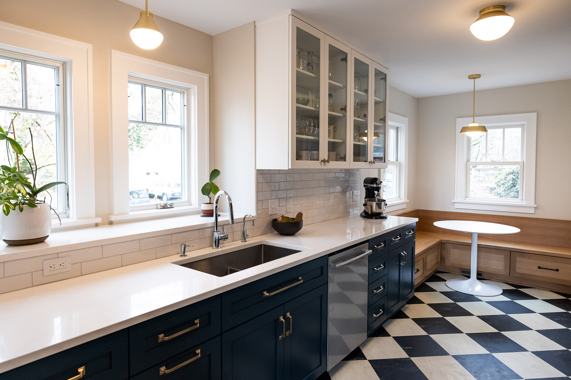 Remodeled kitchen from exterior wall showing deep window sill with plants, navy lower cabinets, glass-front upper cabinets, checkerboard floor, and breakfast nook beyond, Stanton Street Kitchen by Tweto Architecture