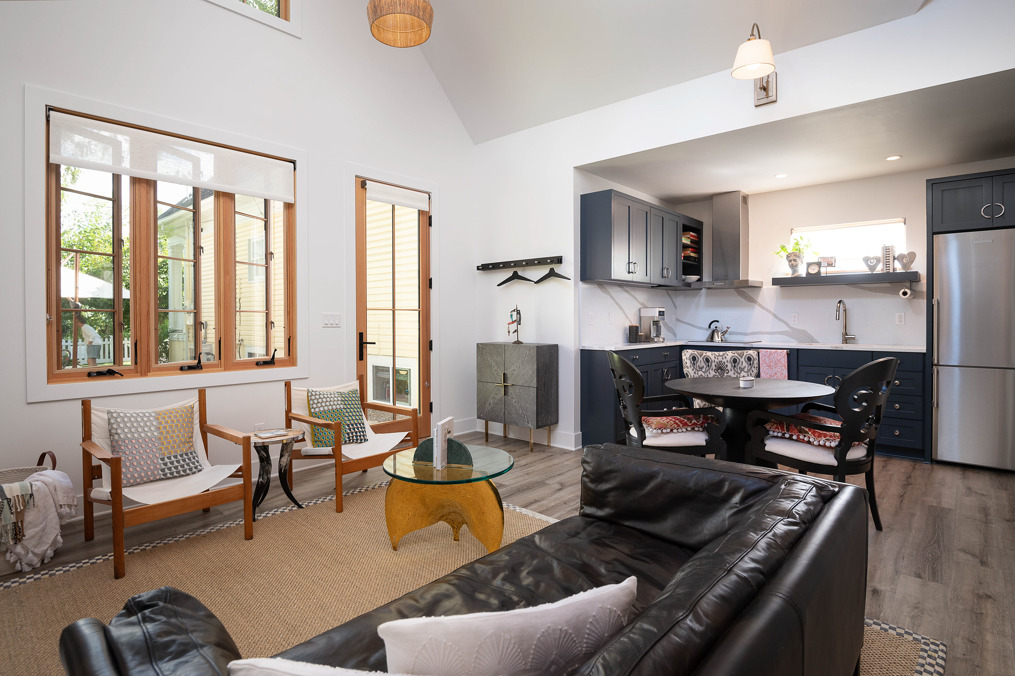 Great room looking toward the yard with large folding wood-framed windows and door, two chairs with colorful pillows, and blue-gray kitchen cabinets visible at right, Vancouver Avenue ADU by Tweto Architecture
