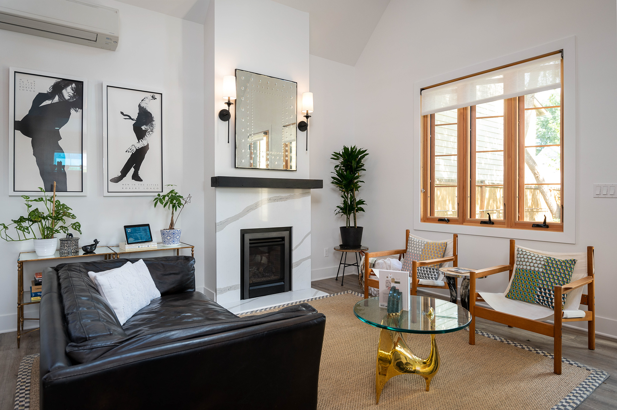 Living room with fireplace, black leather sofa, brass and glass coffee table, two wood chairs, art prints, and large wood-framed window, Vancouver Avenue ADU by Tweto Architecture