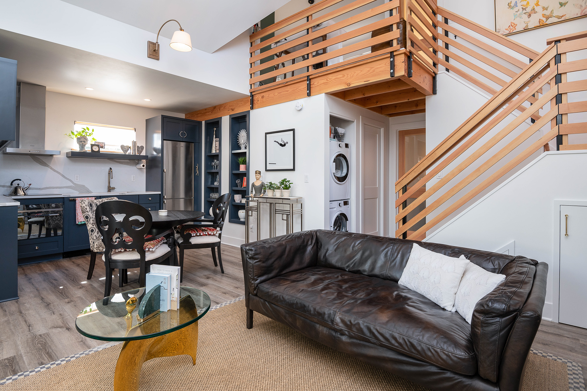 Great room from entry end looking toward kitchen and stair, with leather sofa, glass coffee table, blue-gray kitchen cabinets, and loft railing visible above, Vancouver Avenue ADU by Tweto Architecture