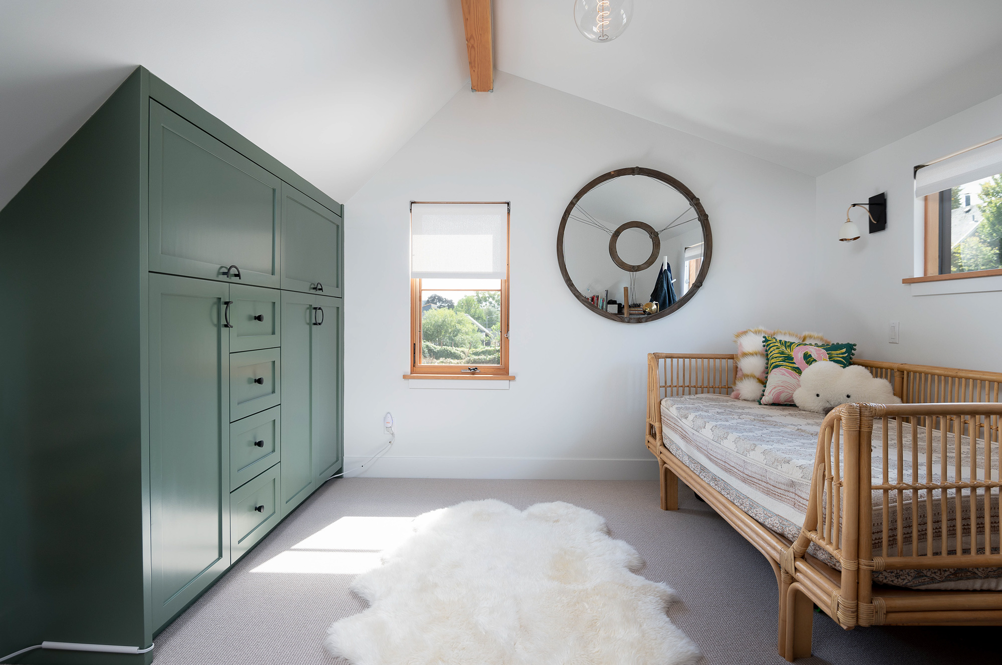 Loft bedroom under the vaulted gable ceiling, with large sage green built-in cabinetry, rattan daybed, round wood mirror, and small wood-framed windows, Vancouver Avenue ADU by Tweto Architecture