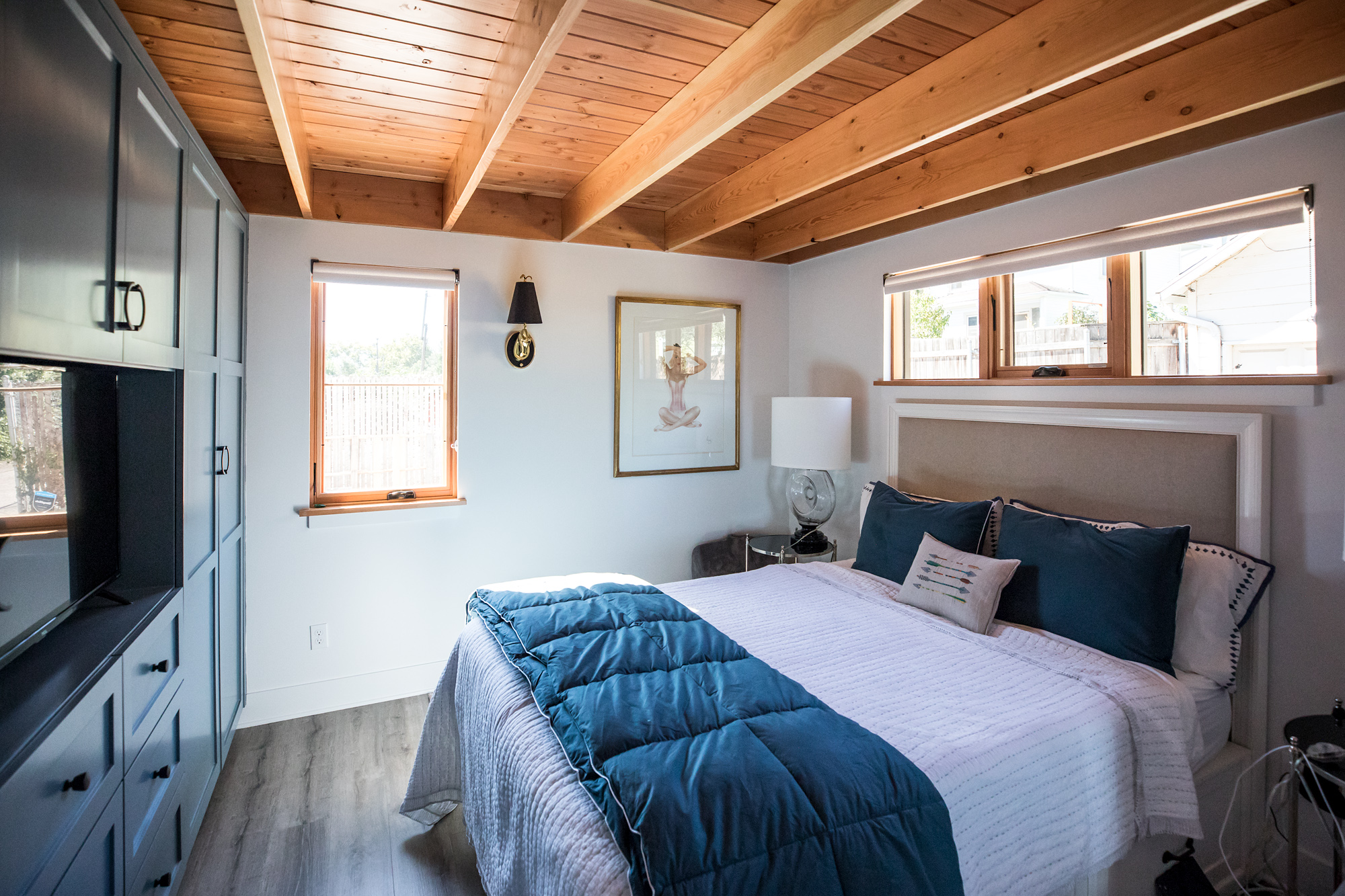 Ground floor bedroom with exposed wood rafter ceiling, blue-gray built-in cabinetry, bed with blue comforter, and clerestory windows, Vancouver Avenue ADU by Tweto Architecture