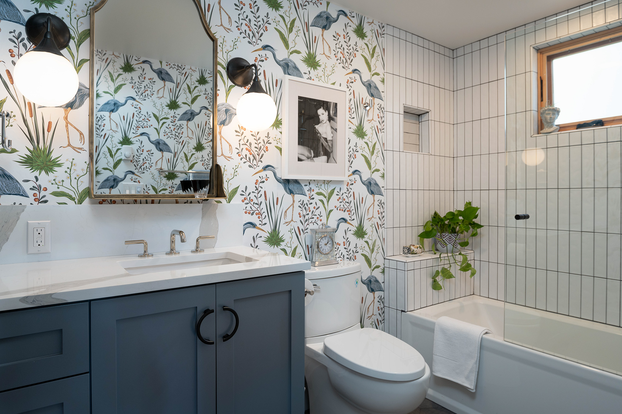 Bathroom with blue-gray vanity and white quartz top, egret botanical wallpaper, gold-framed mirror with globe sconces, and white subway tile tub and shower, Vancouver Avenue ADU by Tweto Architecture